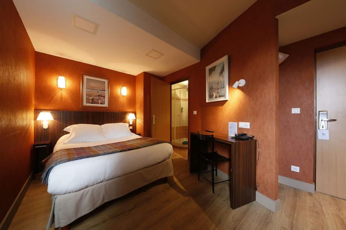 A hotel room with a bed, desk, and chair, a wooden floor, and a door.