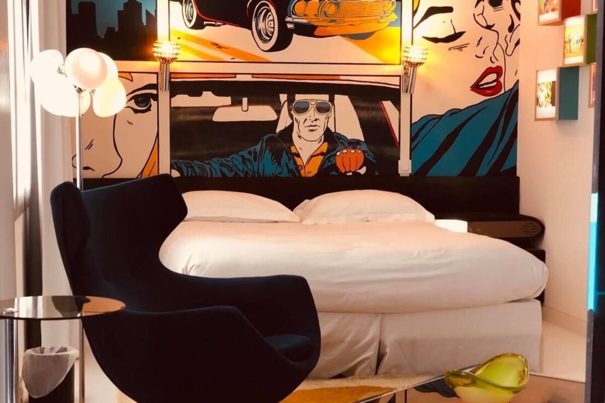 A modern bedroom with a bed, a dark armchair, a glass coffee table, and a cityscape mural on the wall.