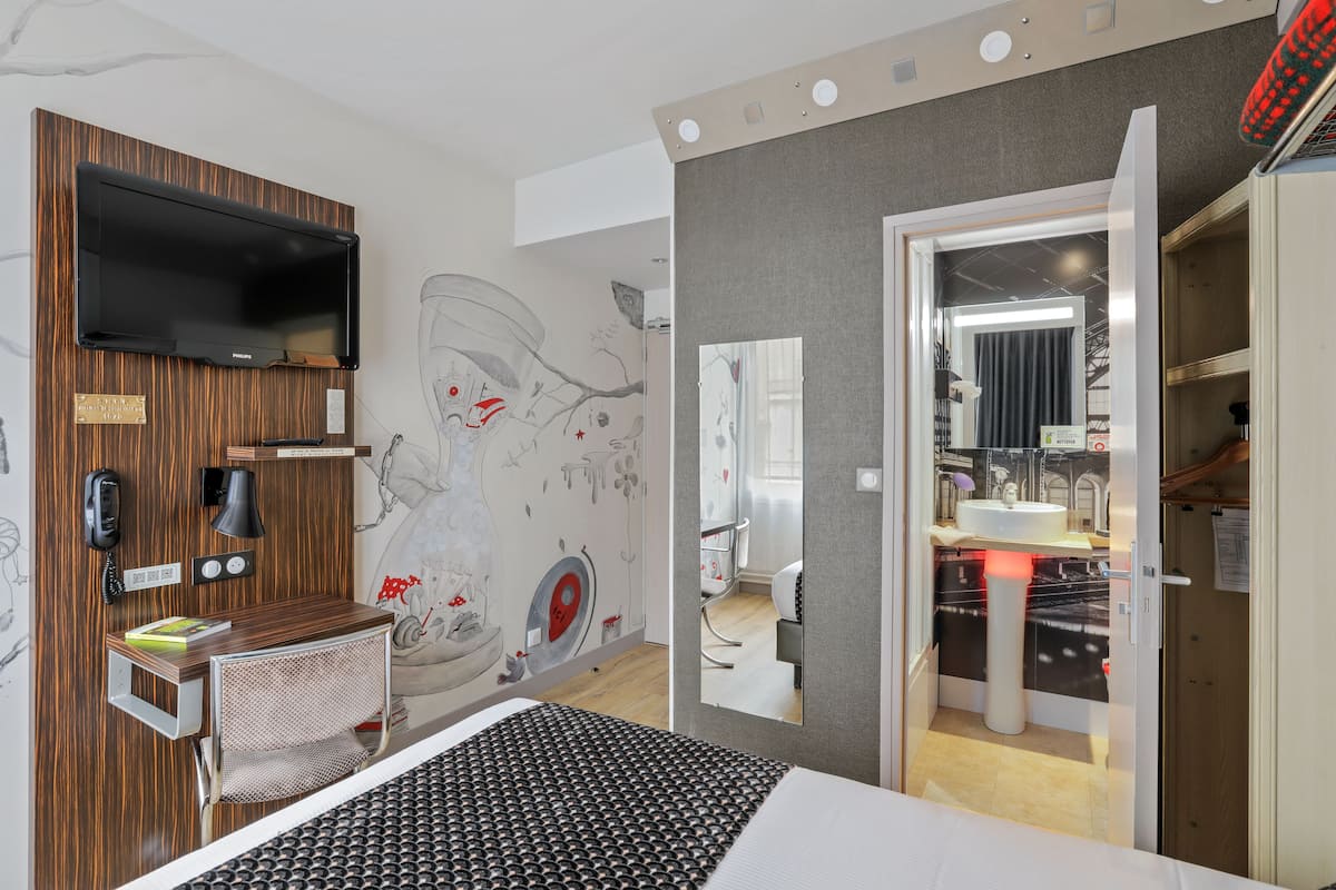 A modern hotel room with a bed, a desk, a chair, a TV, a mirror, and a bathroom visible through an open door.