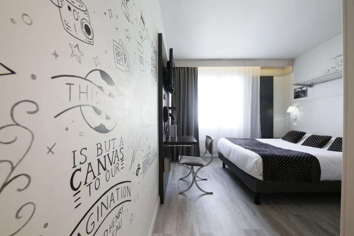 A modern hotel room with a bed, a desk, and a chair. There is a wall with artistic drawings and text.