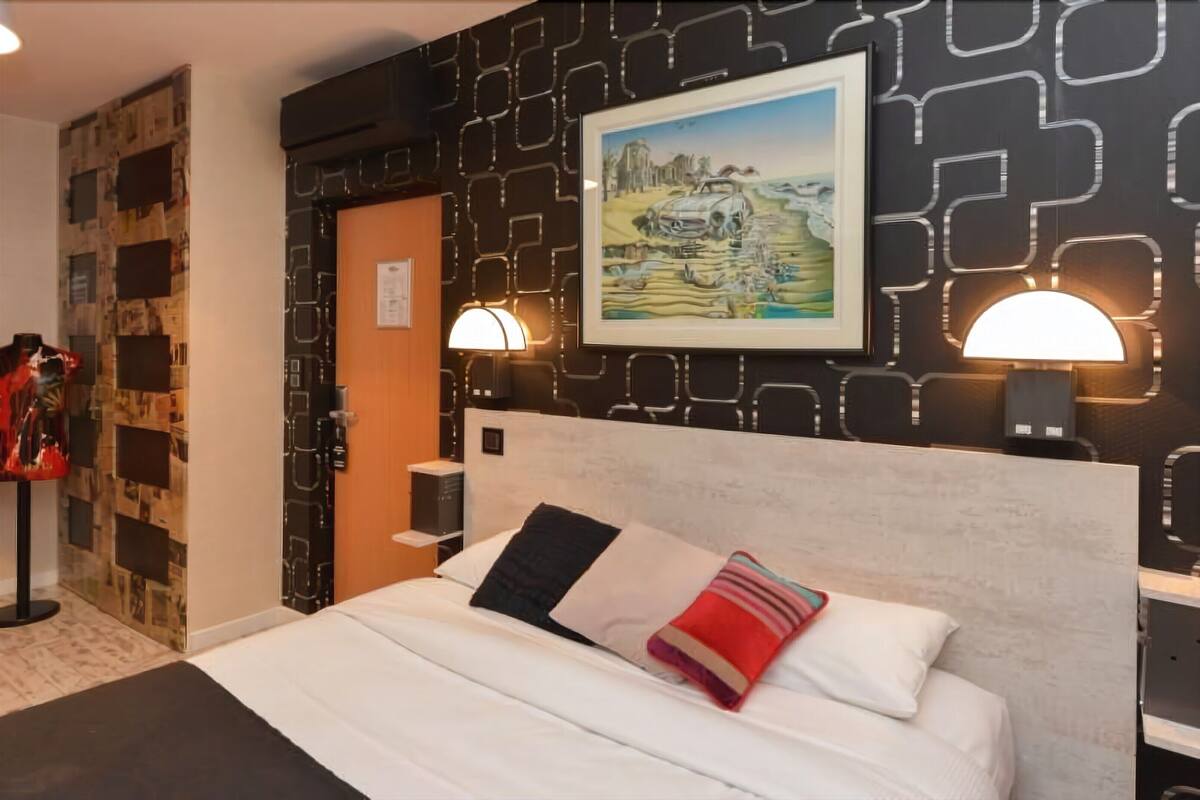 A hotel room with a bed, two wall-mounted lamps, a framed painting, and a unique wall design.