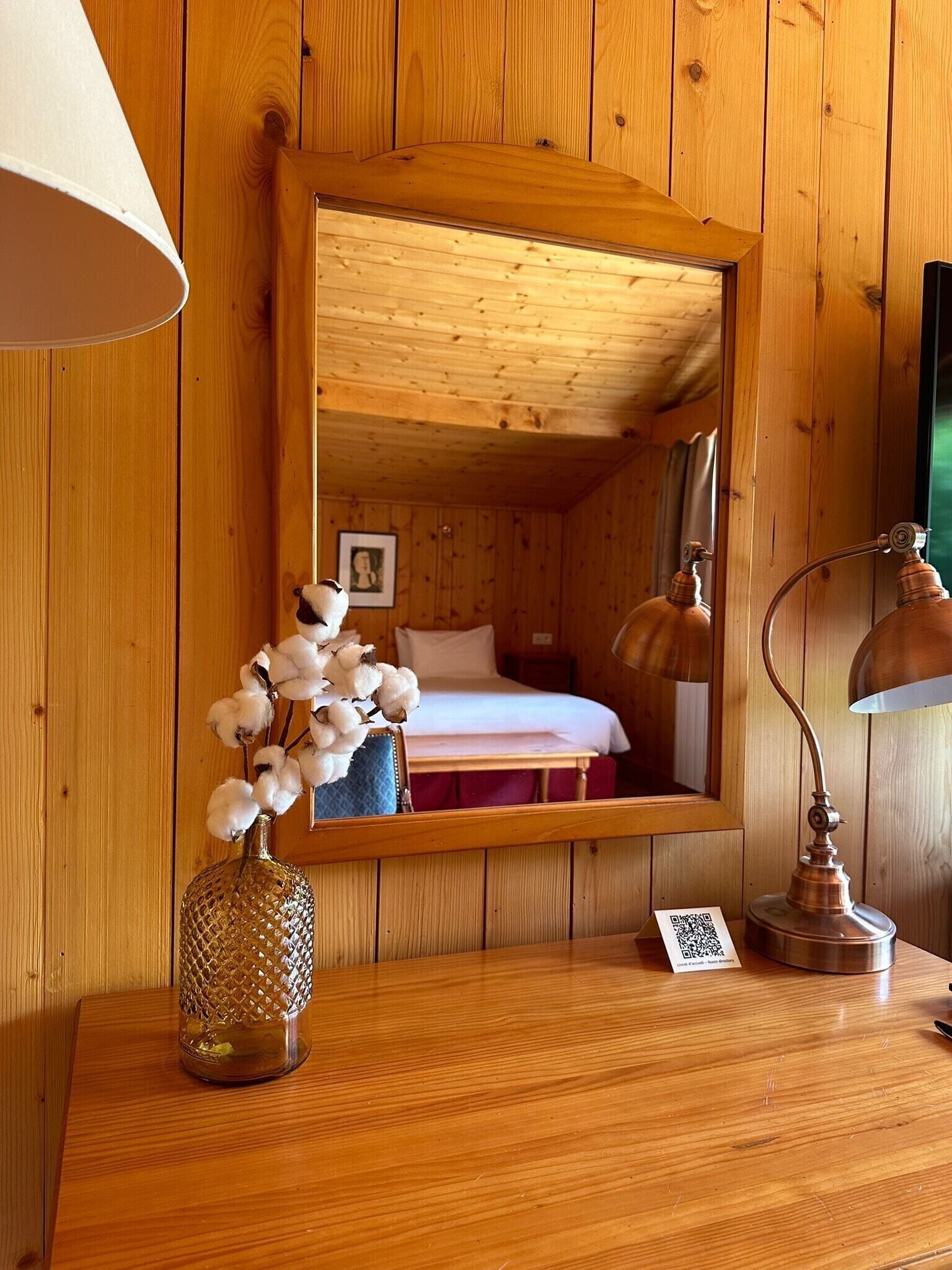Um quarto de madeira com uma cama, um espelho, um abajur e um vaso com algodão.