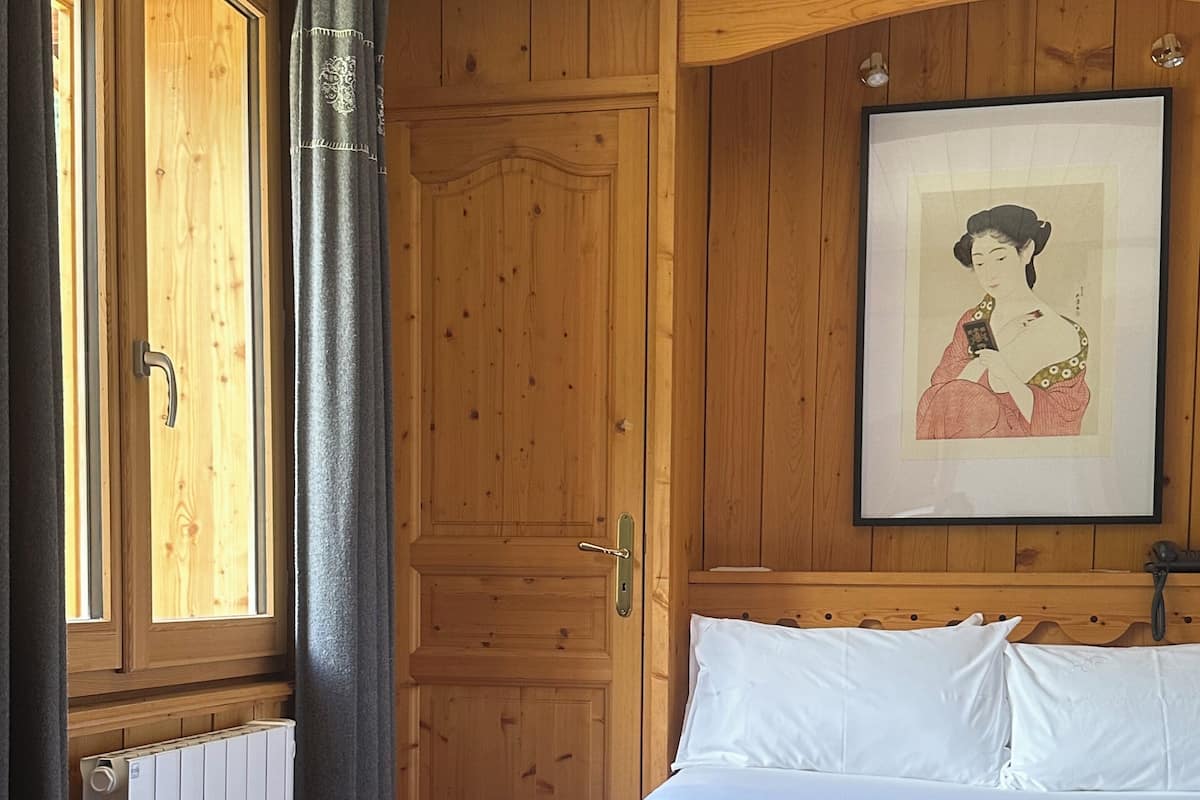 Een kamer met houten lambrisering, een bed, een lijst met een afbeelding aan de muur en een raam met gordijnen.