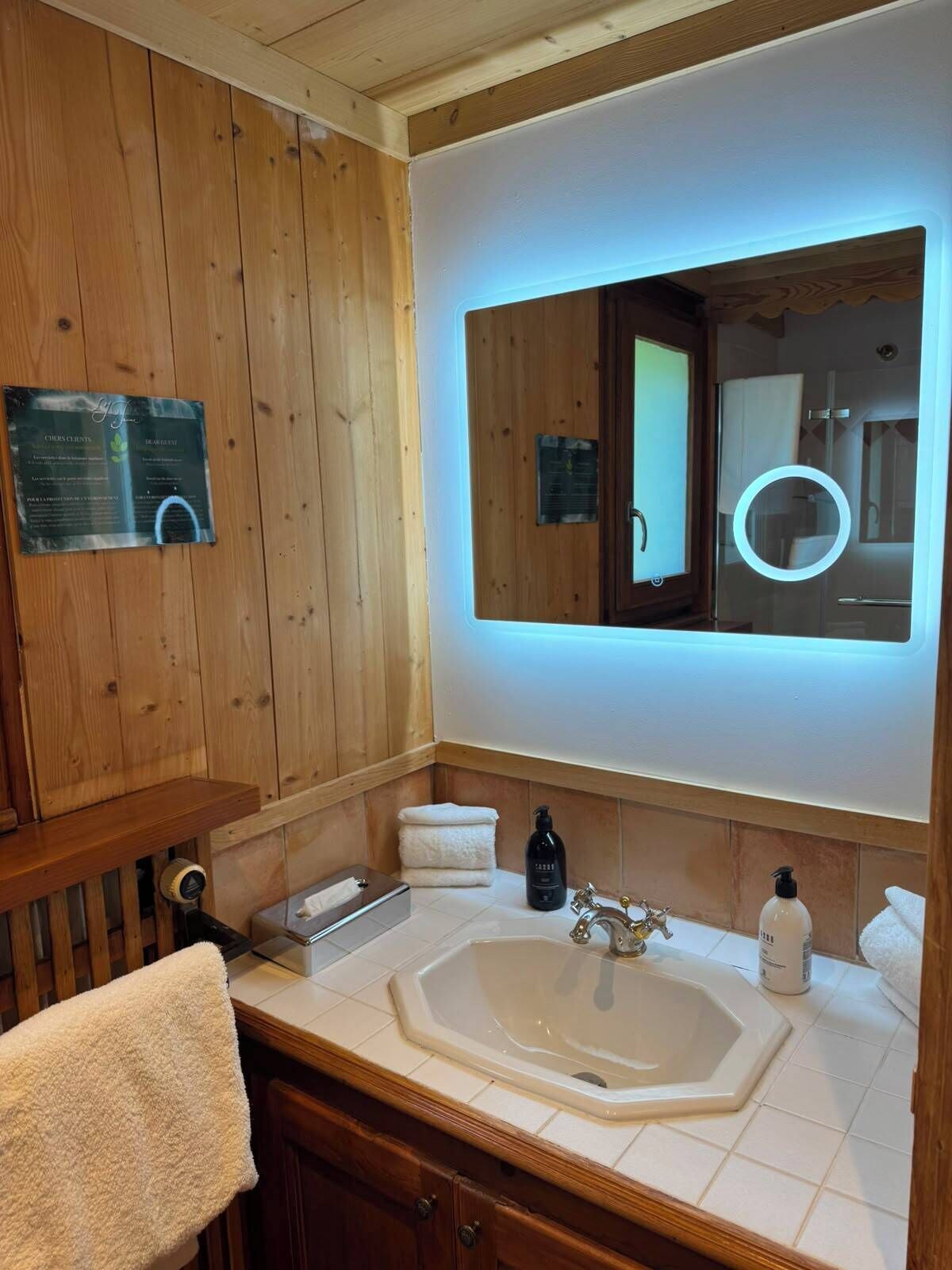 A bathroom with a wooden vanity, a large mirror with a built-in light, a bathtub, and a sink.
