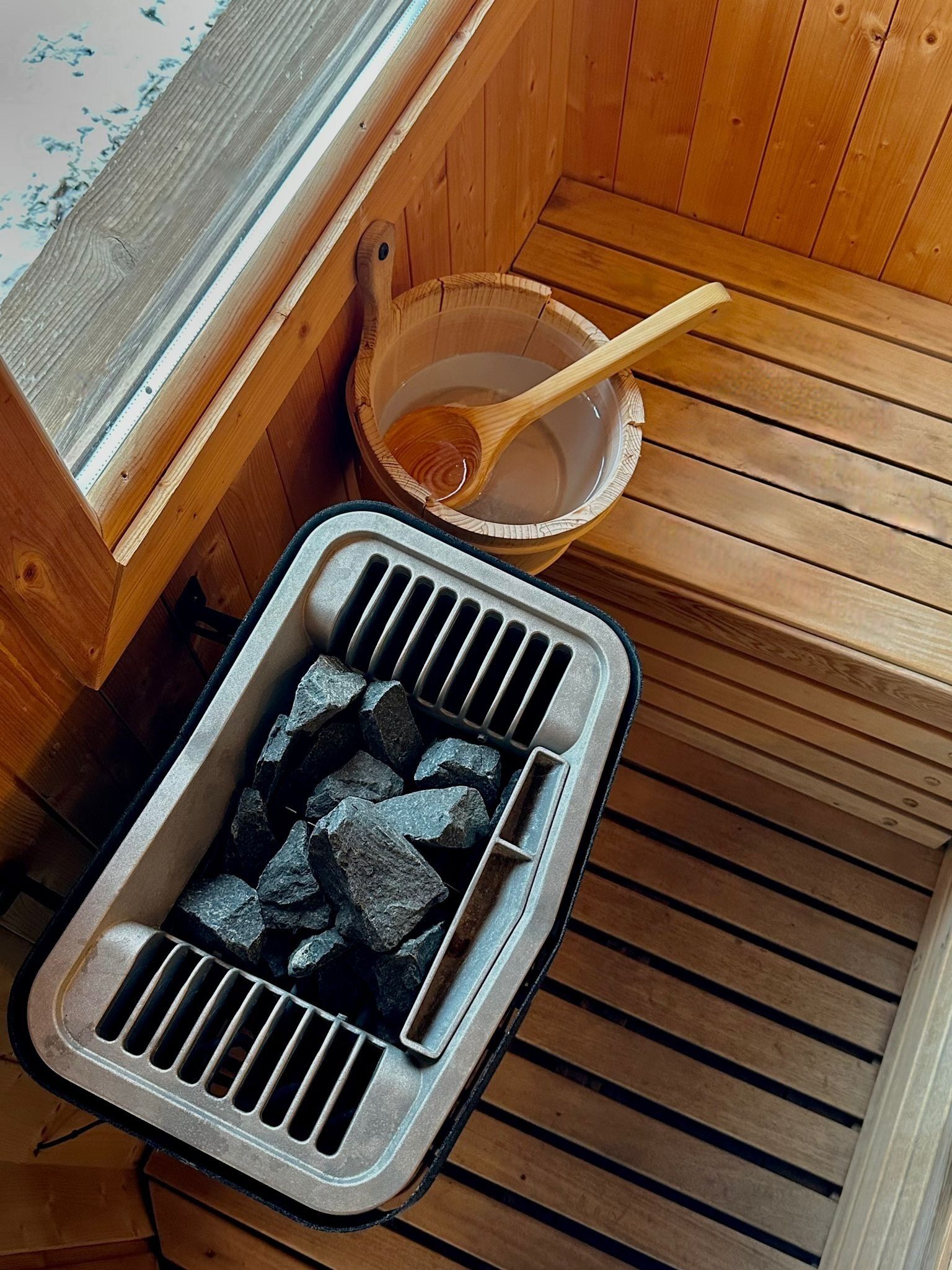 Una sauna con un banco de madera, un cuenco de madera con una cuchara y un recipiente metálico con carbón.