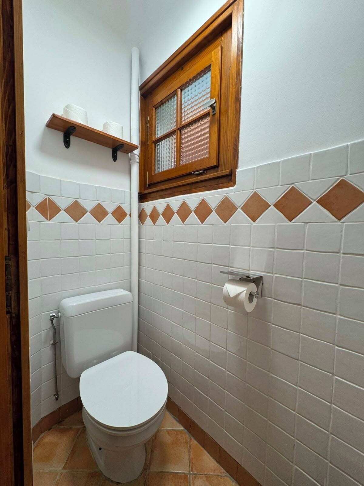 Un baño pequeño con inodoro, ventana y repisa con un rollo de papel higiénico.