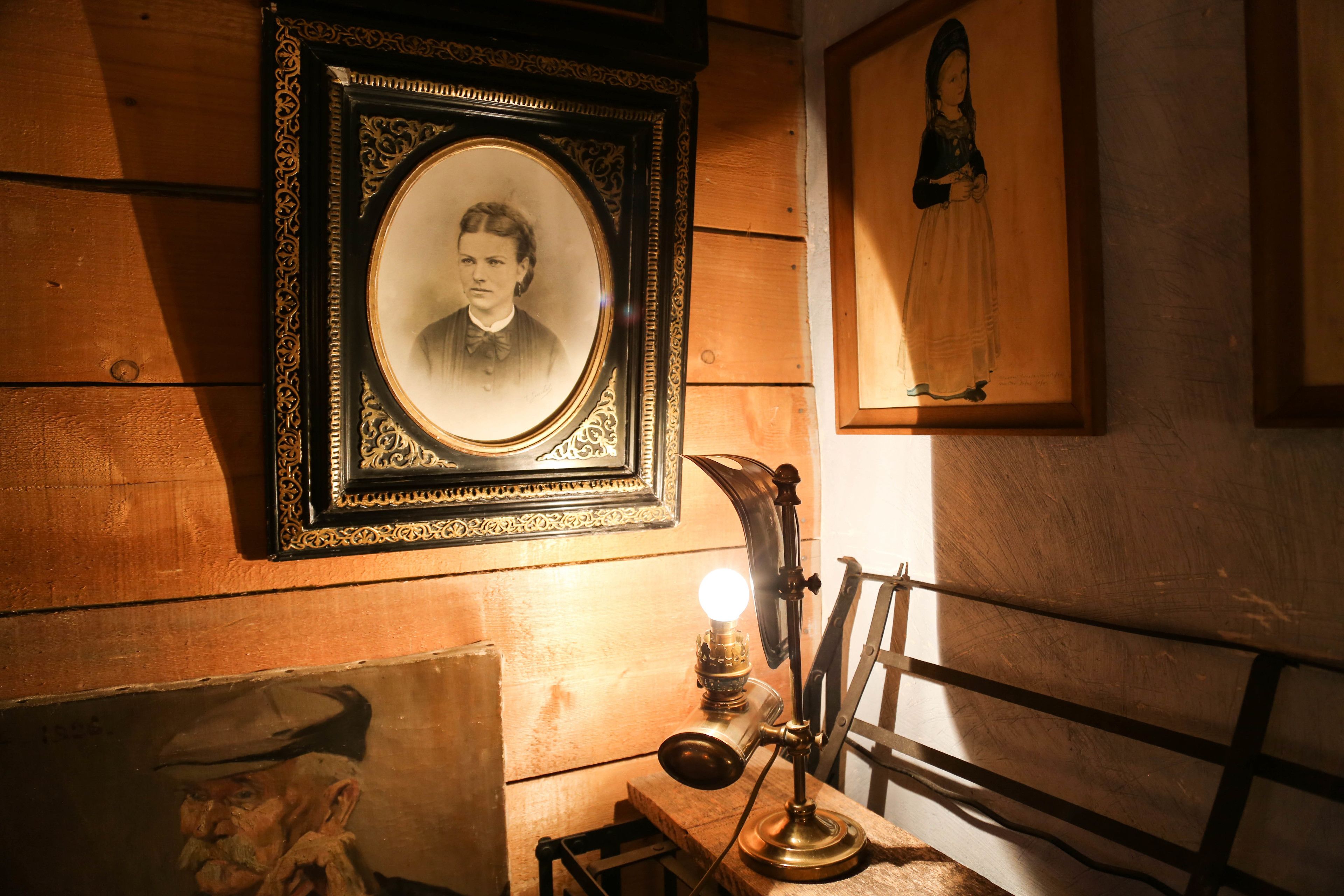 A room with wooden walls, a framed portrait, a vintage desk lamp, and a painting.