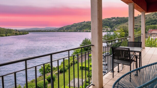 A balcony with a view of a river and hills, a table, and chairs. - Le Beau Rivage (Condrieu)