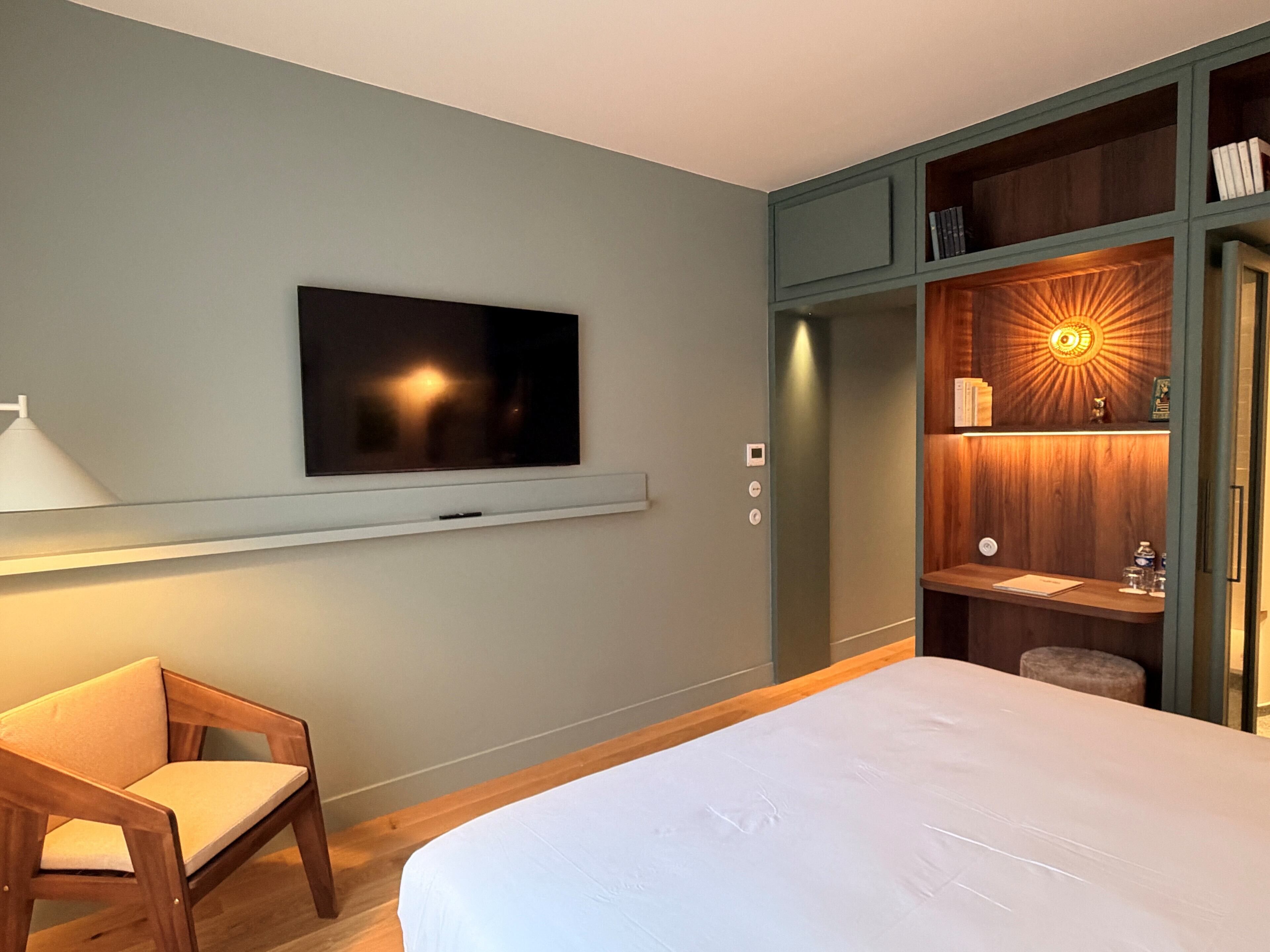 A hotel room with a bed, a TV mounted on the wall, a wooden chair, a built-in bookshelf, and a vanity with a mirror.