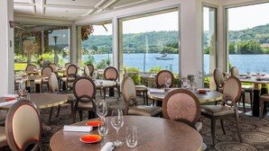A restaurant with tables and chairs arranged for dining, large windows offering a view of a lake and a bridge, and a well-lit interior. - Le Beau Rivage (Condrieu)