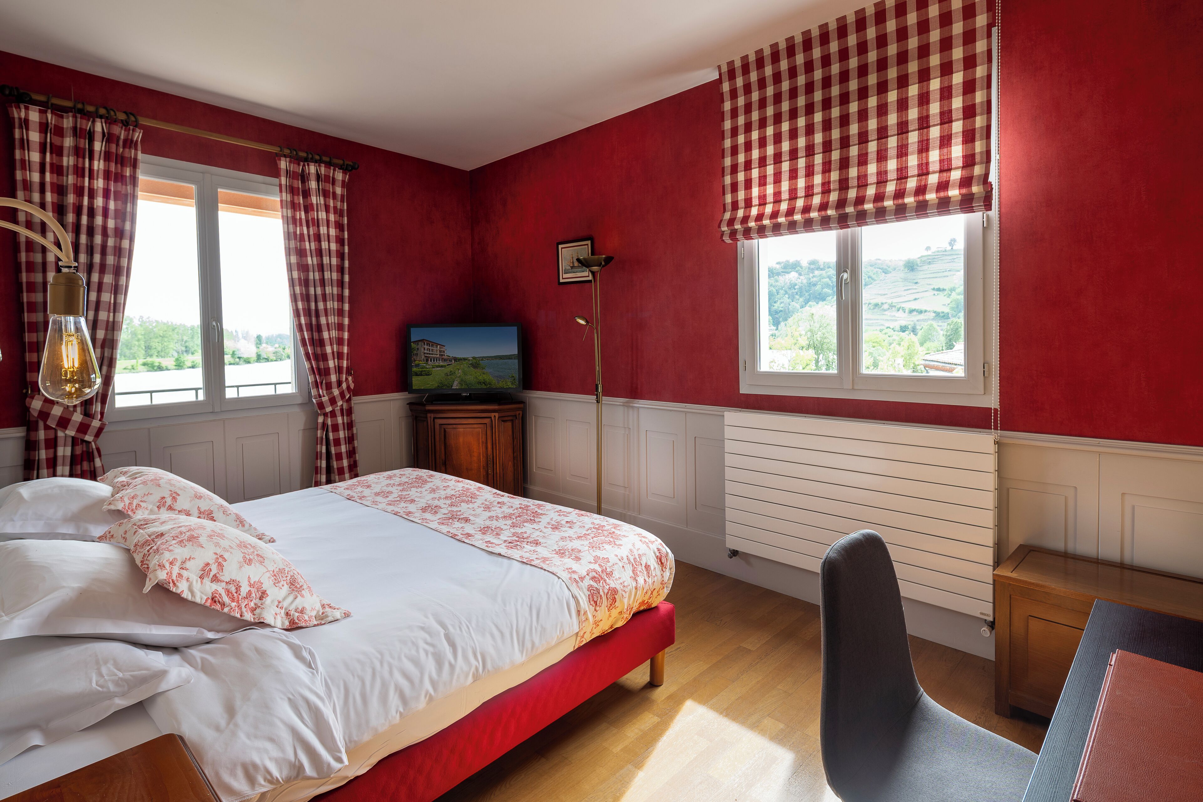 A bedroom with a bed, a desk, a chair, a lamp, and two windows with curtains.