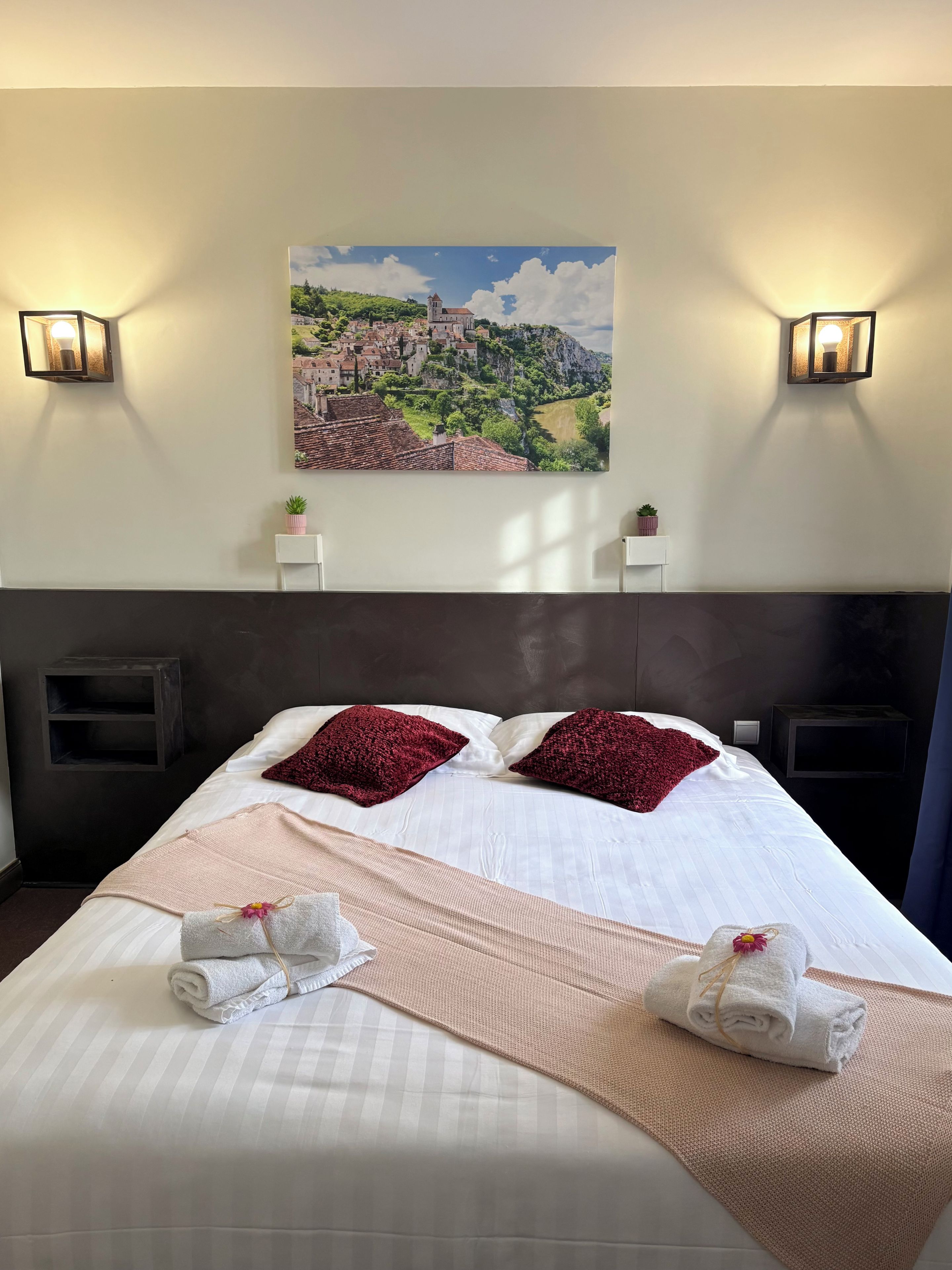 Una camera d'albergo con un letto grande, due cuscini, una testiera, un quadro raffigurante un borgo, lampade da comodino, una piccola pianta e una televisione.