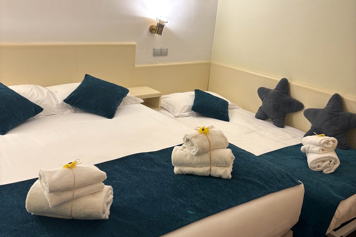 Quarto de hotel com duas camas, cada uma com colchas azul-esverdeadas e lençóis brancos, mesa de cabeceira com abajur e duas molduras na parede.