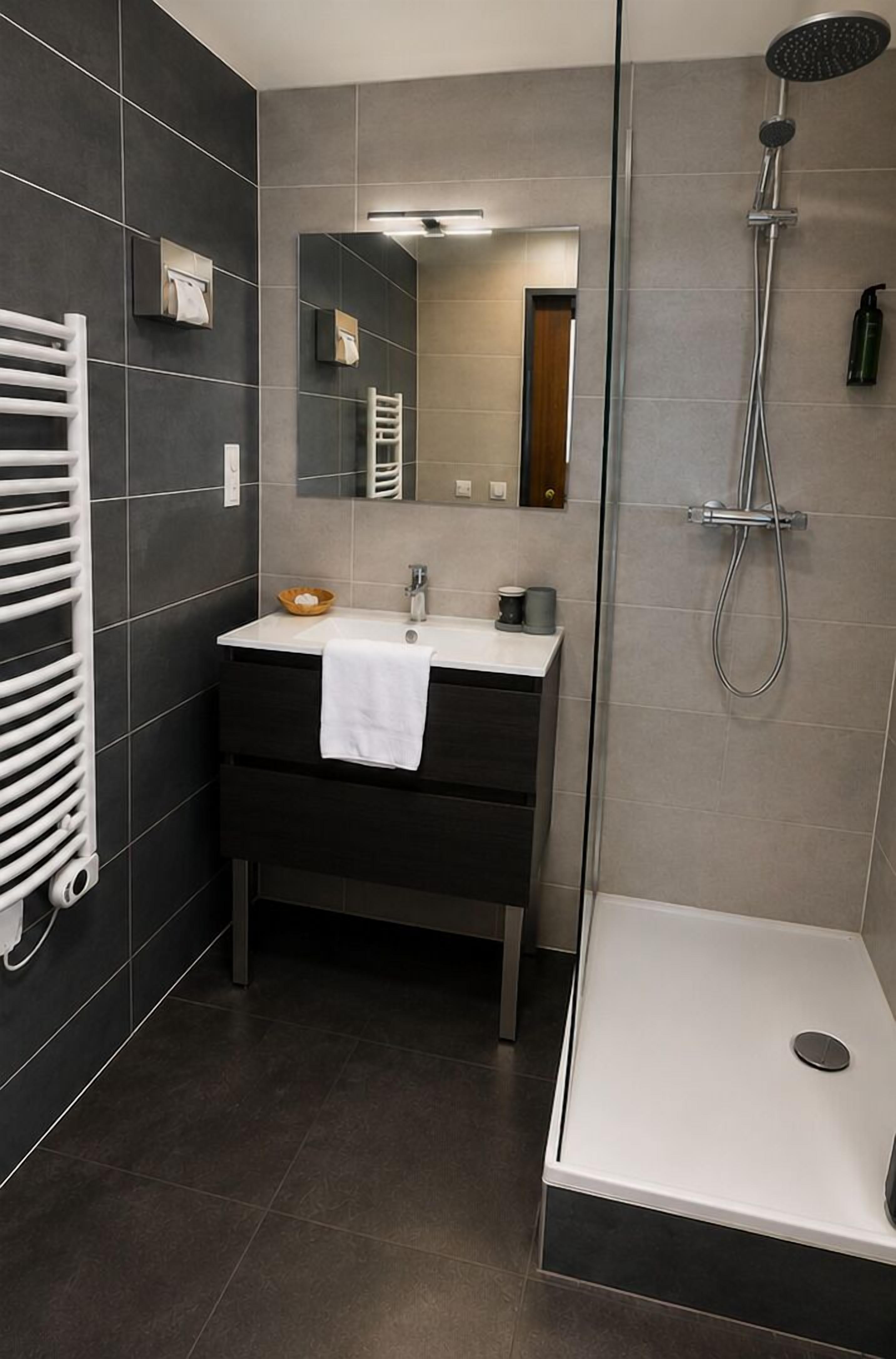 Un baño moderno con lavamanos blanco, baldosas oscuras, ducha y toallero.