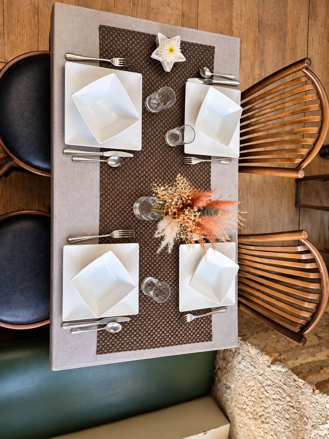A rectangular dining table with four white square plates, silverware, and clear glasses, set on a woven placemat.
