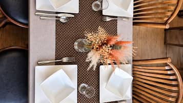 A rectangular dining table with four white square plates, silverware, and clear glasses, set on a woven placemat.