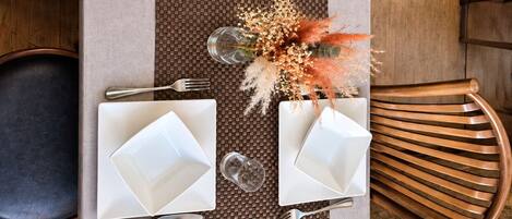 A rectangular dining table with four white square plates, silverware, and clear glasses, set on a woven placemat.