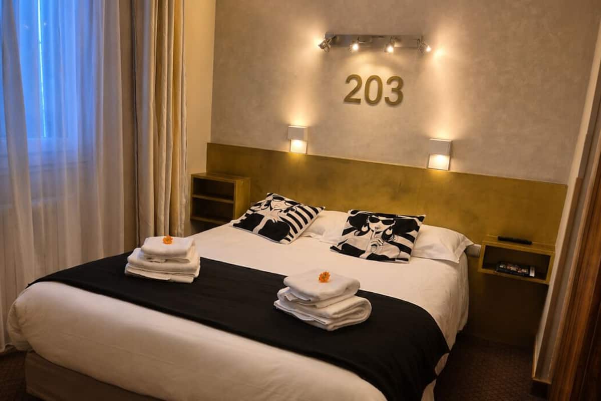 Quarto de hotel com uma cama grande, duas mesas de cabeceira, um número fixado na parede e uma janela com cortinas.