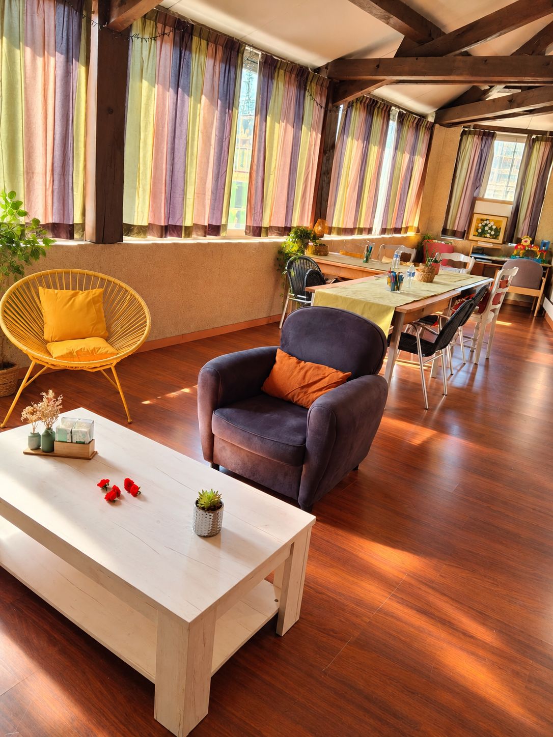 A room with a wooden floor, a white table with a vase and red flowers, a dark armchair with an orange pillow, and a dining area with a table and chairs.