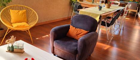 Una habitación con piso de madera, una mesa blanca con un florero y flores rojas, un sillón oscuro con un cojín naranja, y un comedor con una mesa y sillas.