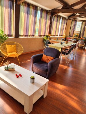 A room with a wooden floor, a white table with a vase and red flowers, a dark armchair with an orange pillow, and a dining area with a table and chairs. - Le Lion d'Or (Gramat)