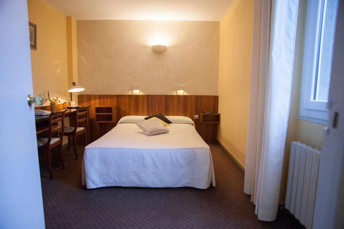 A hotel room with a bed, bedside tables, a desk with a chair, a lamp, and a window with curtains.