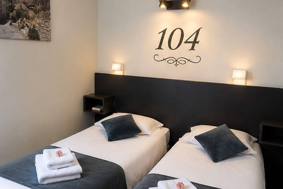 Quarto de hotel com duas camas, um número na parede e uma mesinha de cabeceira.