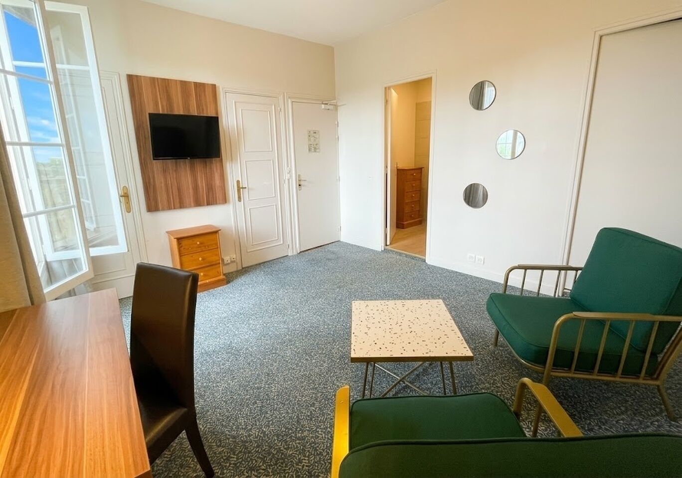 Una habitación con una mesa de madera, dos sillas, una mesita, un televisor, un armario y una puerta que da a otra estancia.