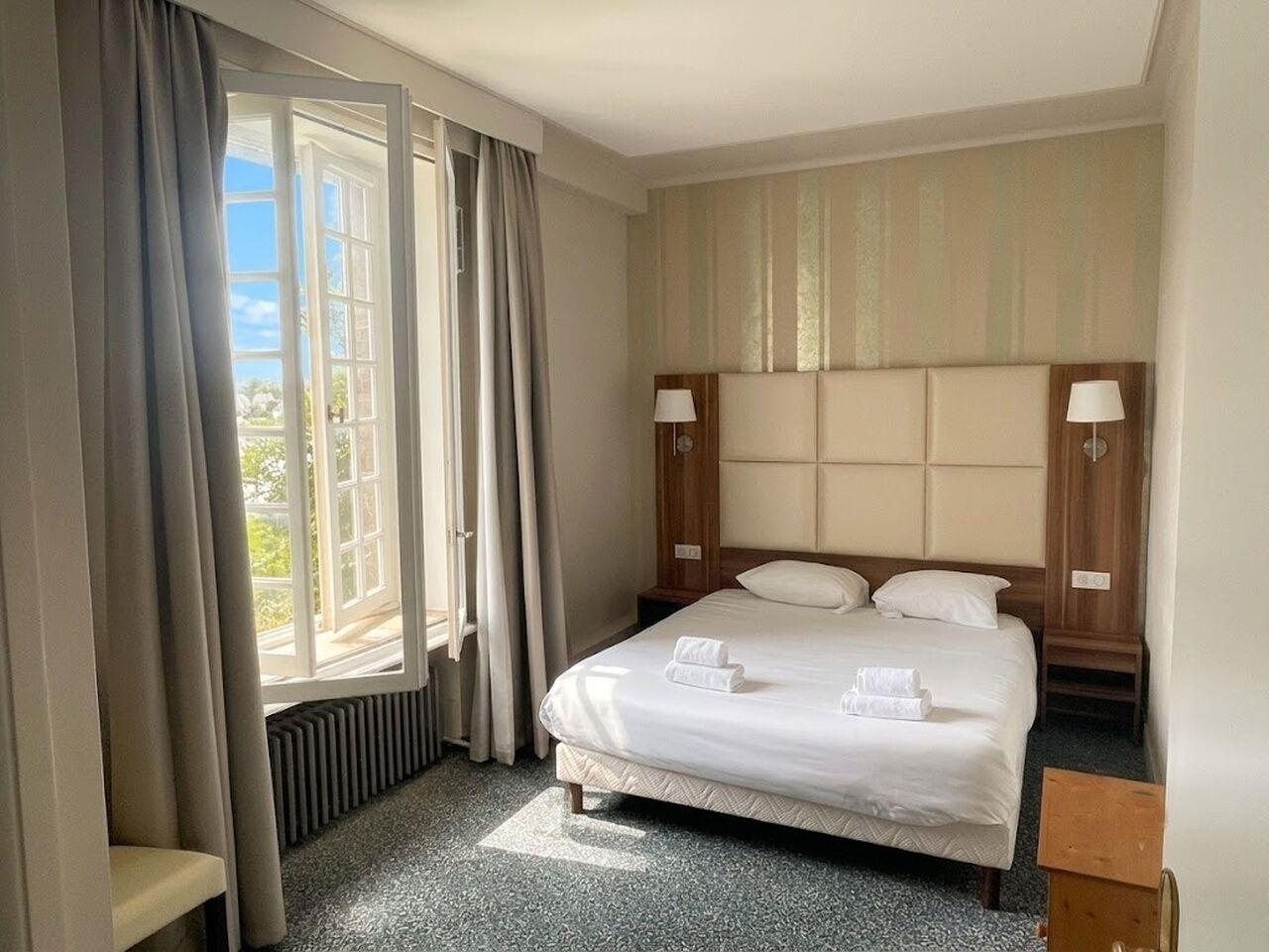 A hotel room with a bed, two bedside tables, a window with curtains, and a view of the outdoors.