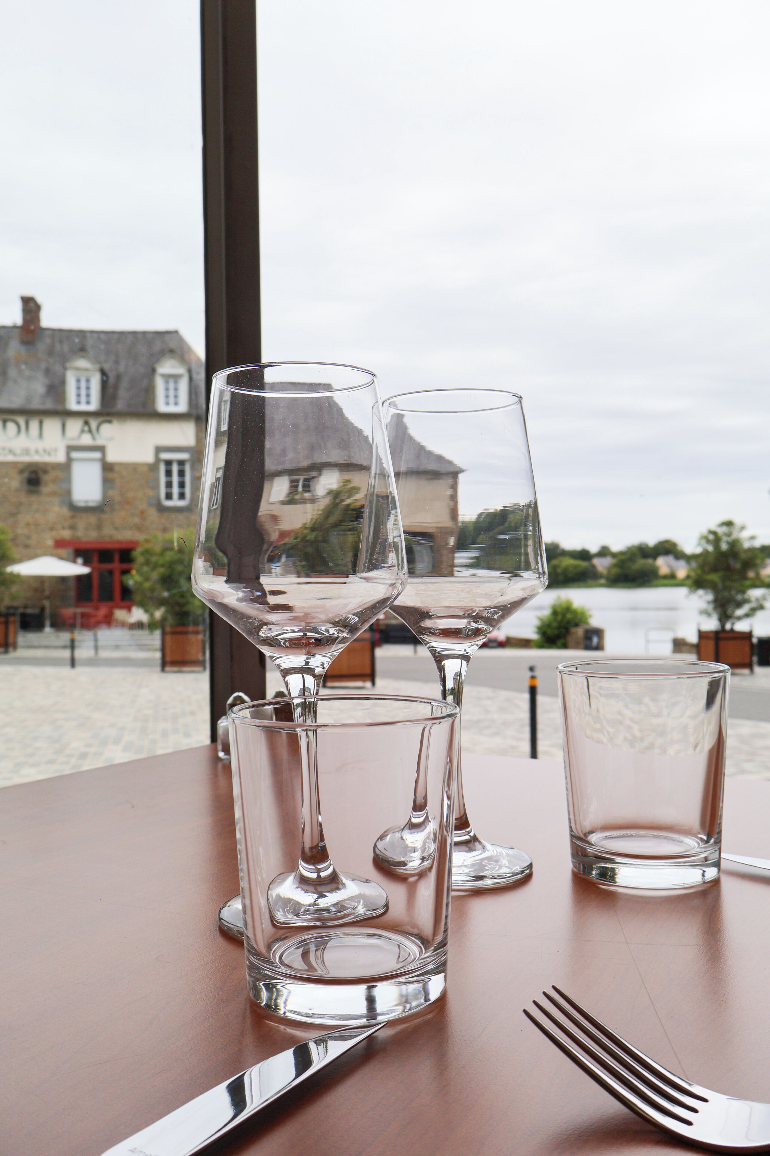 Una mesa con tres copas de vino, un tenedor, un cuchillo y una cuchara, con vista a un edificio y una masa de agua al fondo.