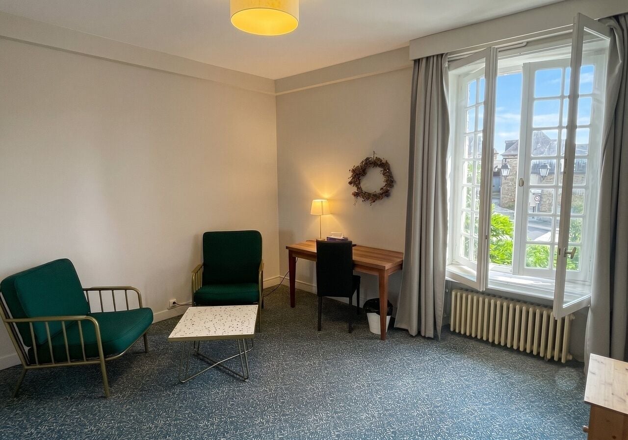 A room with a desk, two chairs, a lamp, a radiator, and a window with curtains.