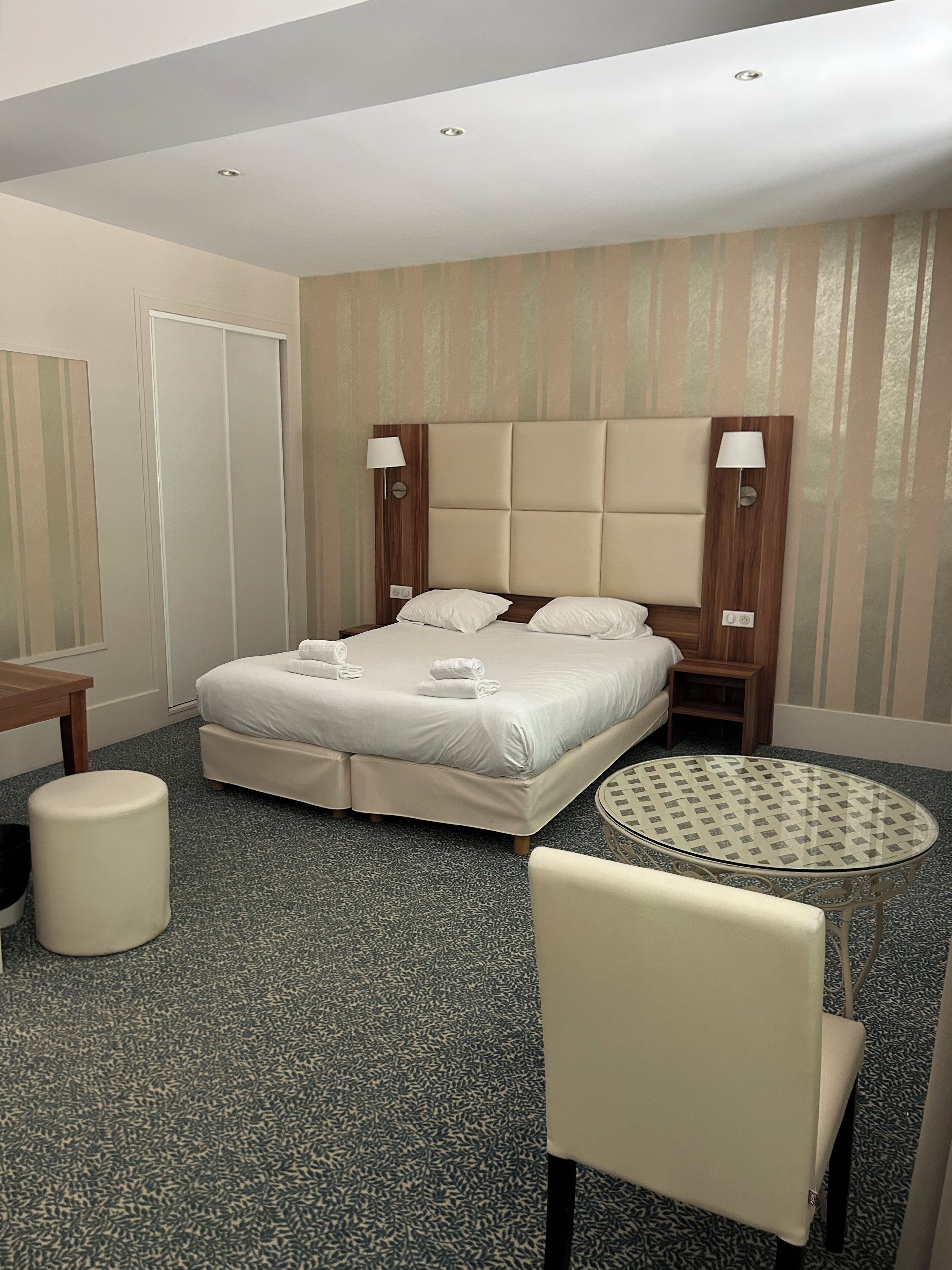 A hotel room with a bed, a round table, a chair, a desk, and a lamp.