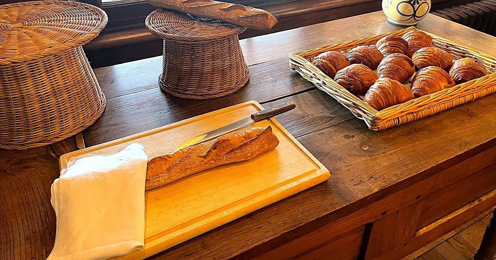 Un comptoir en bois sur lequel se trouve un panier de croissants, un autre panier en osier, ainsi qu’une planche à découper en bois avec une baguette.