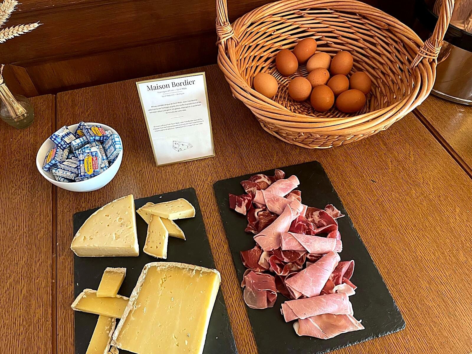 Una mesa de madera con una pizarra negra que muestra queso y embutidos, una cesta de mimbre con huevos, y un bol de aperitivos envasados.