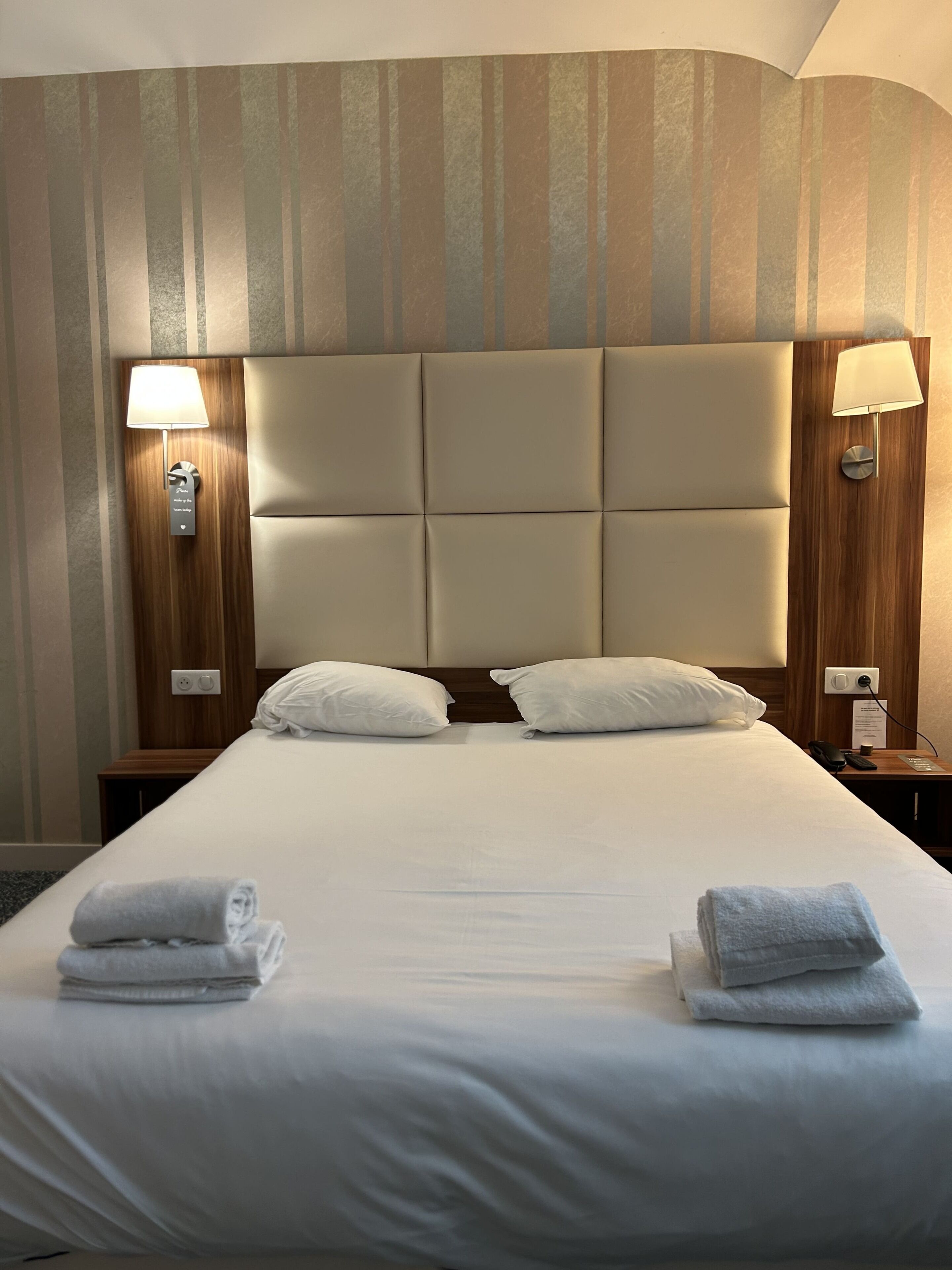 A hotel room with a large bed, two bedside lamps, a wooden headboard, and a wall with striped wallpaper.