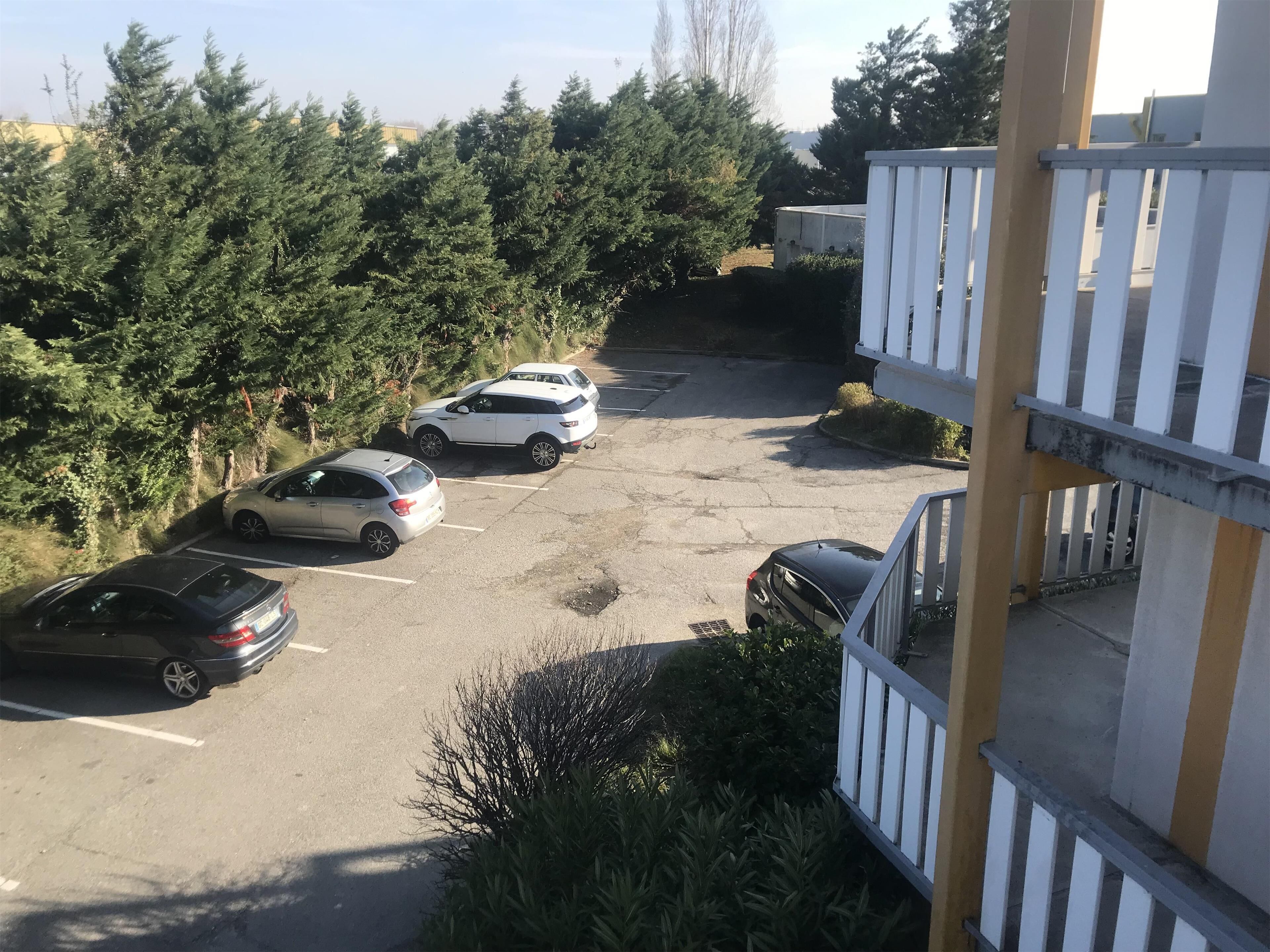 Un estacionamiento con varios autos, rodeado de árboles y un edificio con barandilla blanca.