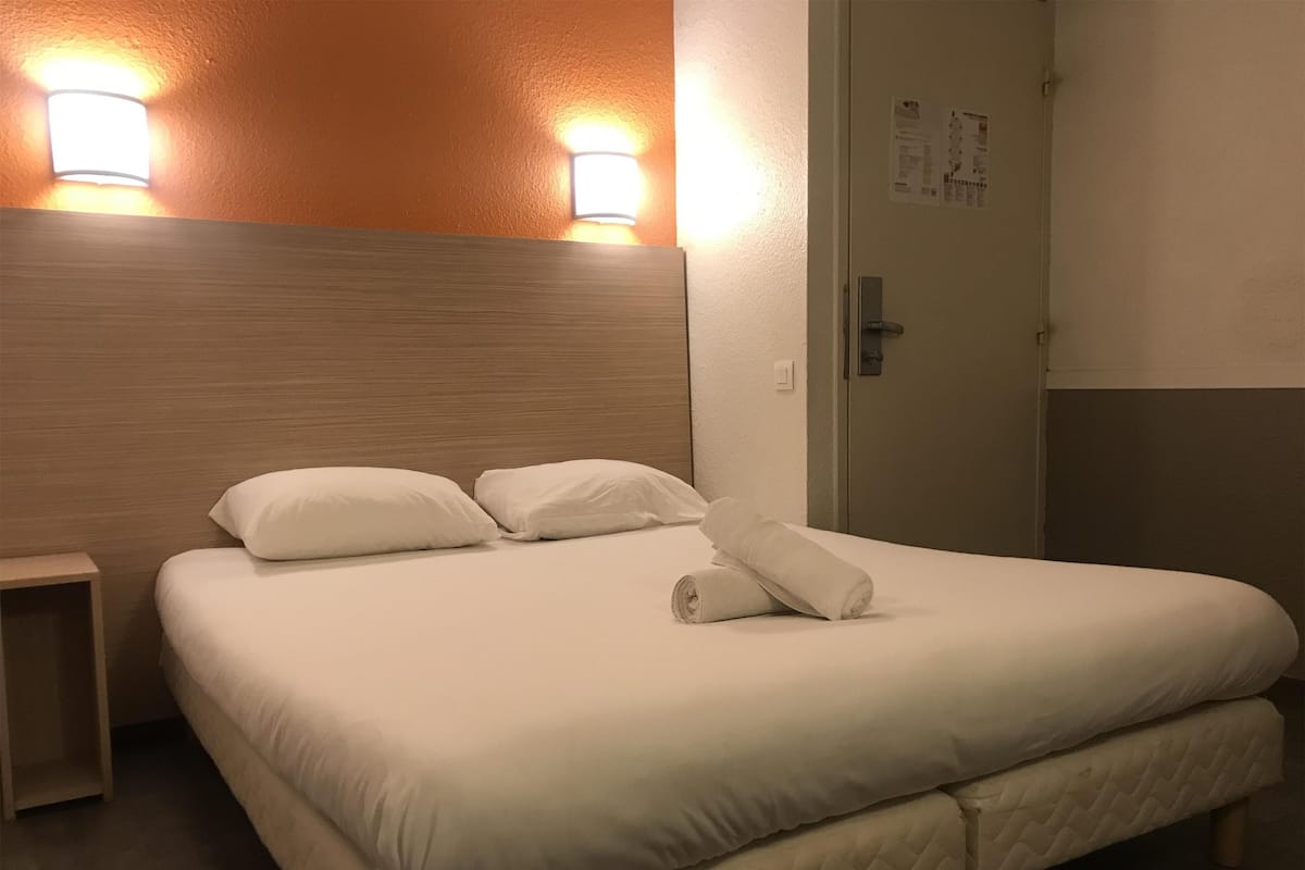 A neatly made bed with white linens and pillows, a wooden headboard, and two wall-mounted lights.