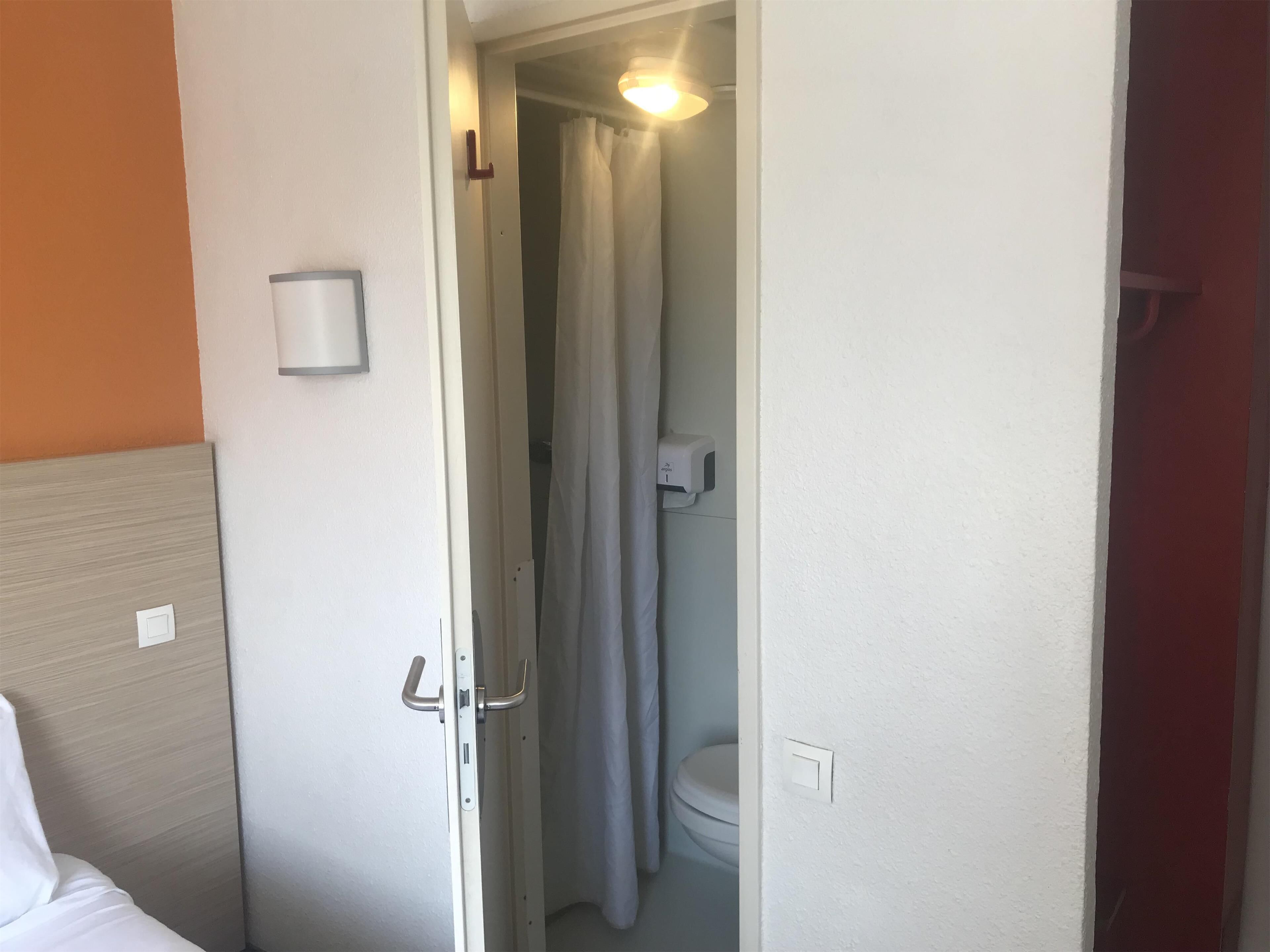 Une chambre d’hôtel avec un lit, une salle de bain avec des toilettes et un sèche-mains, et une porte avec une poignée.