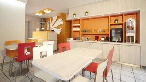 A dining area with a long table and chairs, a kitchen area with shelves, and a refrigerator. - Premiere Classe Avignon Courtine (Avignon)