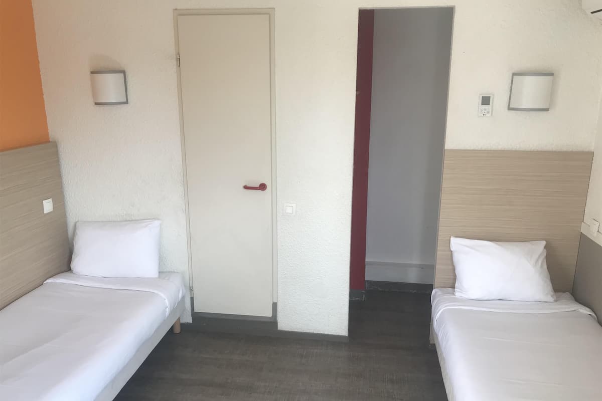 A hotel room with two single beds, a door, a light switch, and a thermostat on the wall.