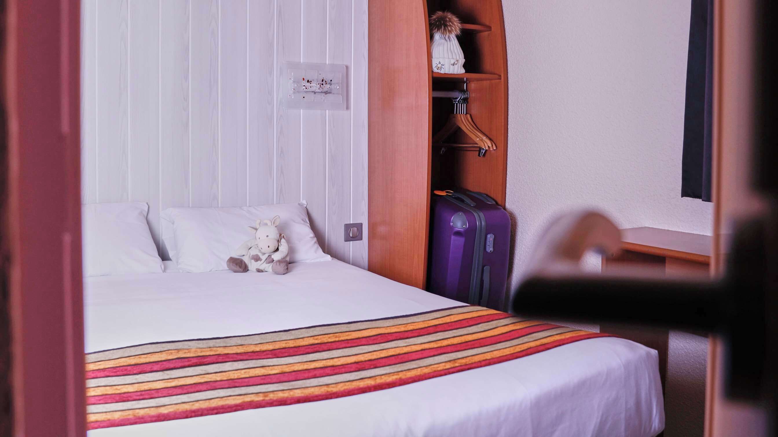 A hotel room with a bed, a striped blanket, a plush toy, a purple suitcase, and a wooden wardrobe.
