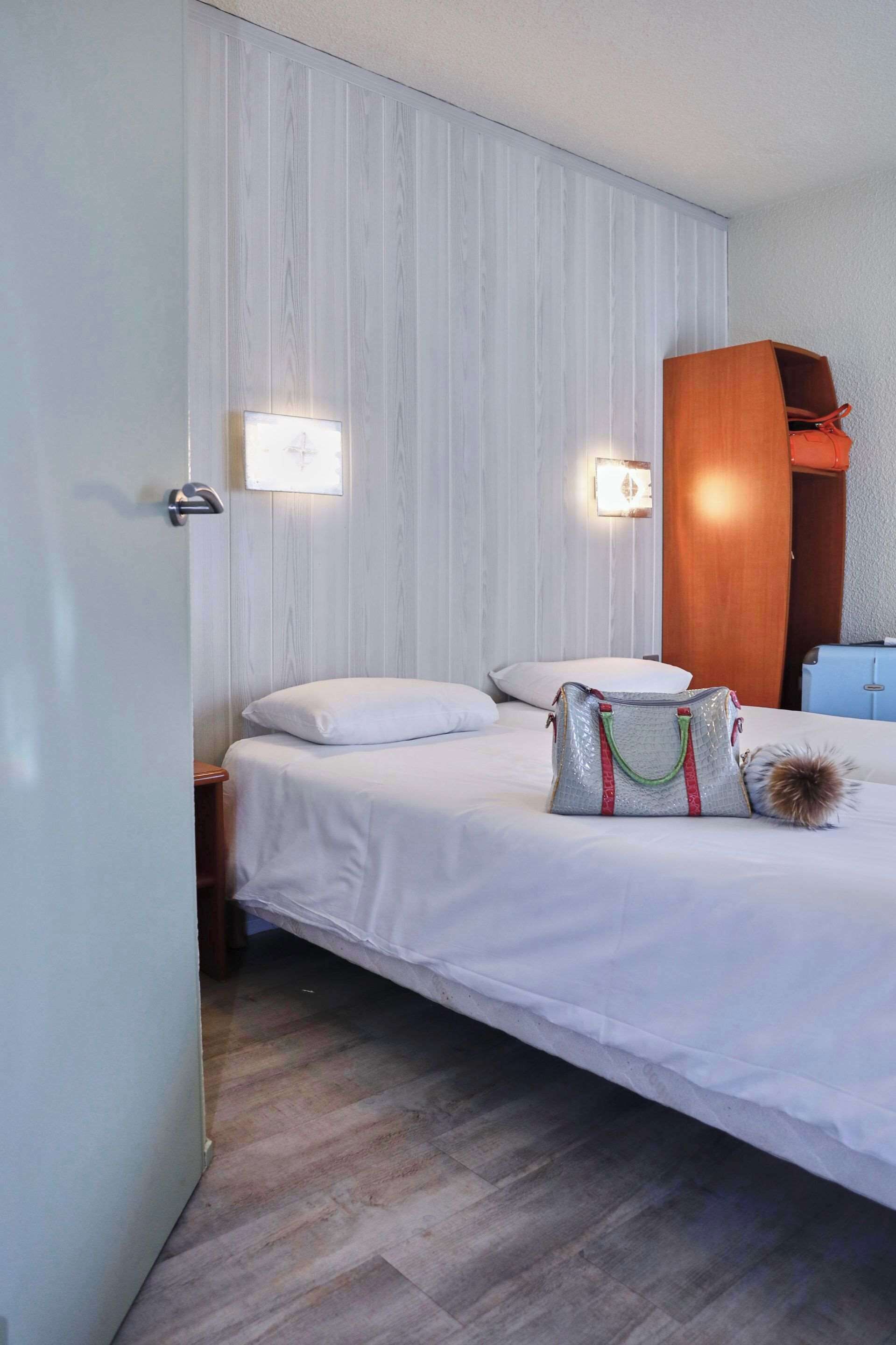 Una habitación de hotel con una cama, lámparas de noche, un armario de madera y una maleta en el suelo.