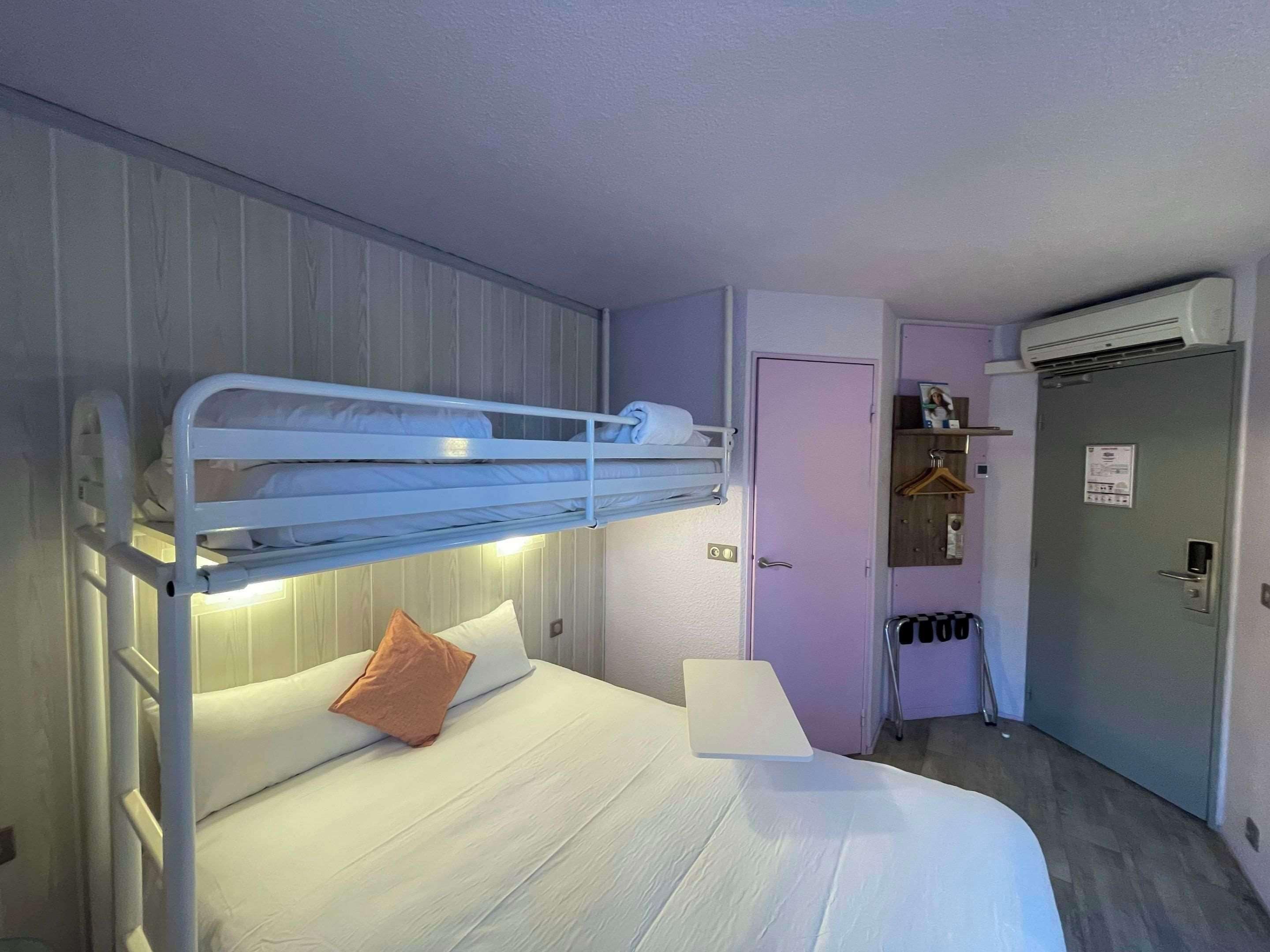 Una cama litera con un colchón individual y una almohada, una mesita de noche, una puerta con un equipo de aire acondicionado, y un pequeño pasillo con un espejo y un perchero.