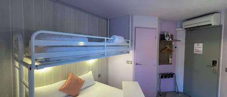 Una cama litera con un colchón individual y una almohada, una mesita de noche, una puerta con un equipo de aire acondicionado, y un pequeño pasillo con un espejo y un perchero.