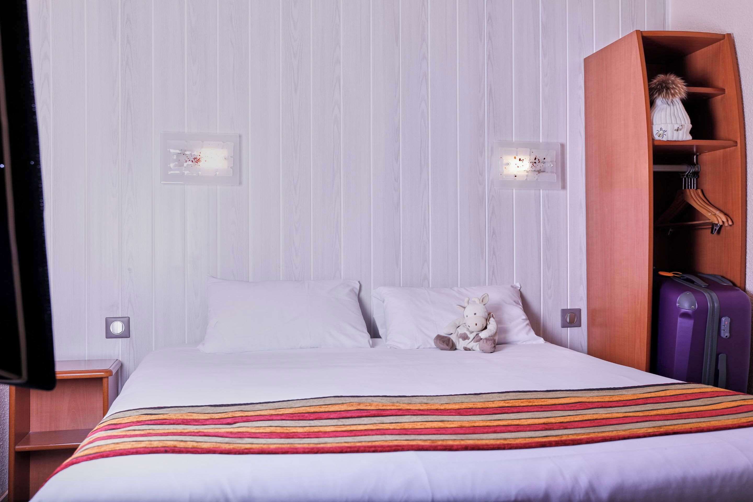 Une chambre d’hôtel avec un lit, une couverture à rayures, un petit animal en peluche, une armoire en bois et une valise violette.