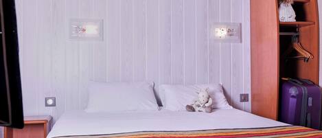 Une chambre d’hôtel avec un lit, une couverture à rayures, un petit animal en peluche, une armoire en bois et une valise violette.