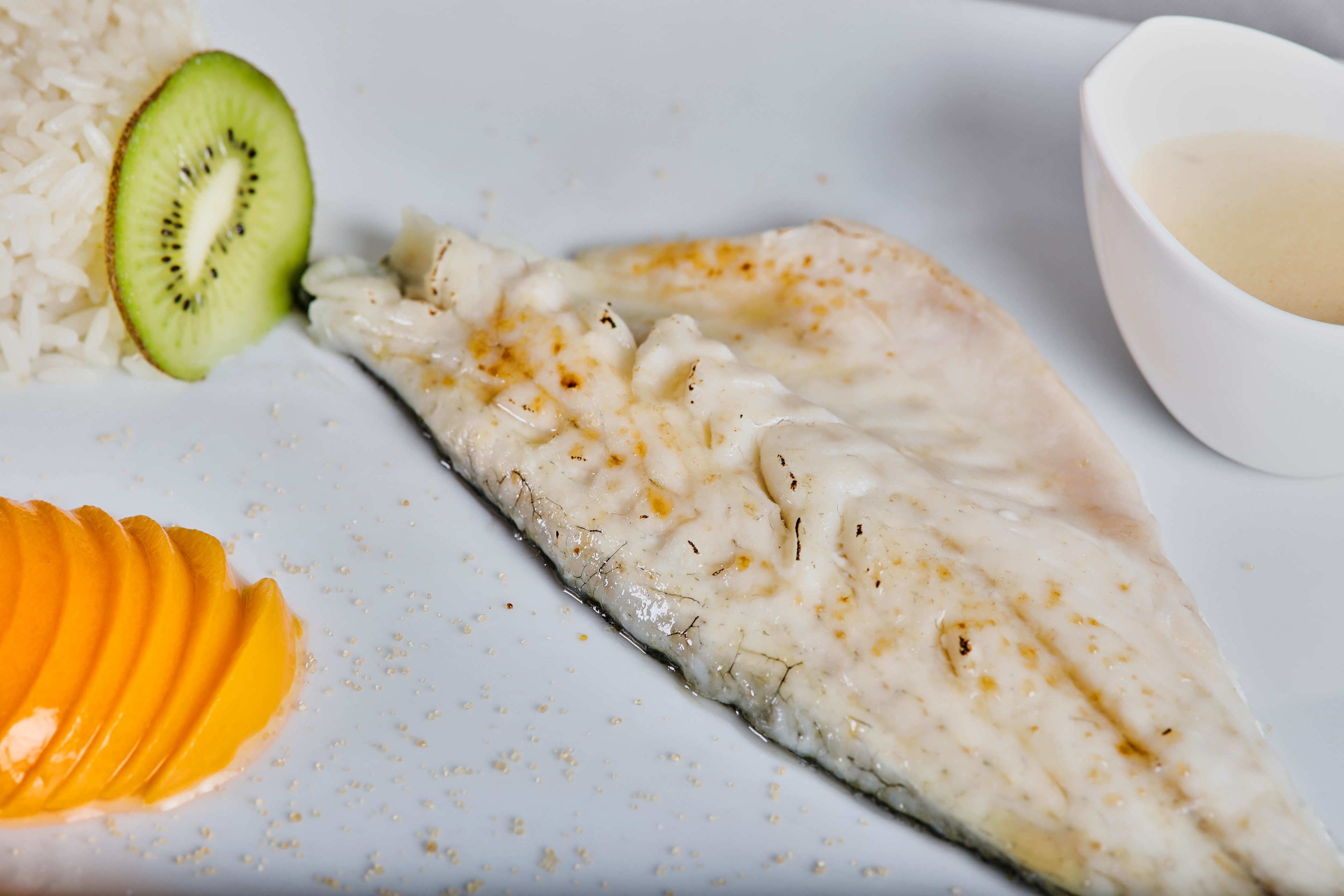 A white plate with a fish fillet, a slice of mango, and a small bowl of sauce.