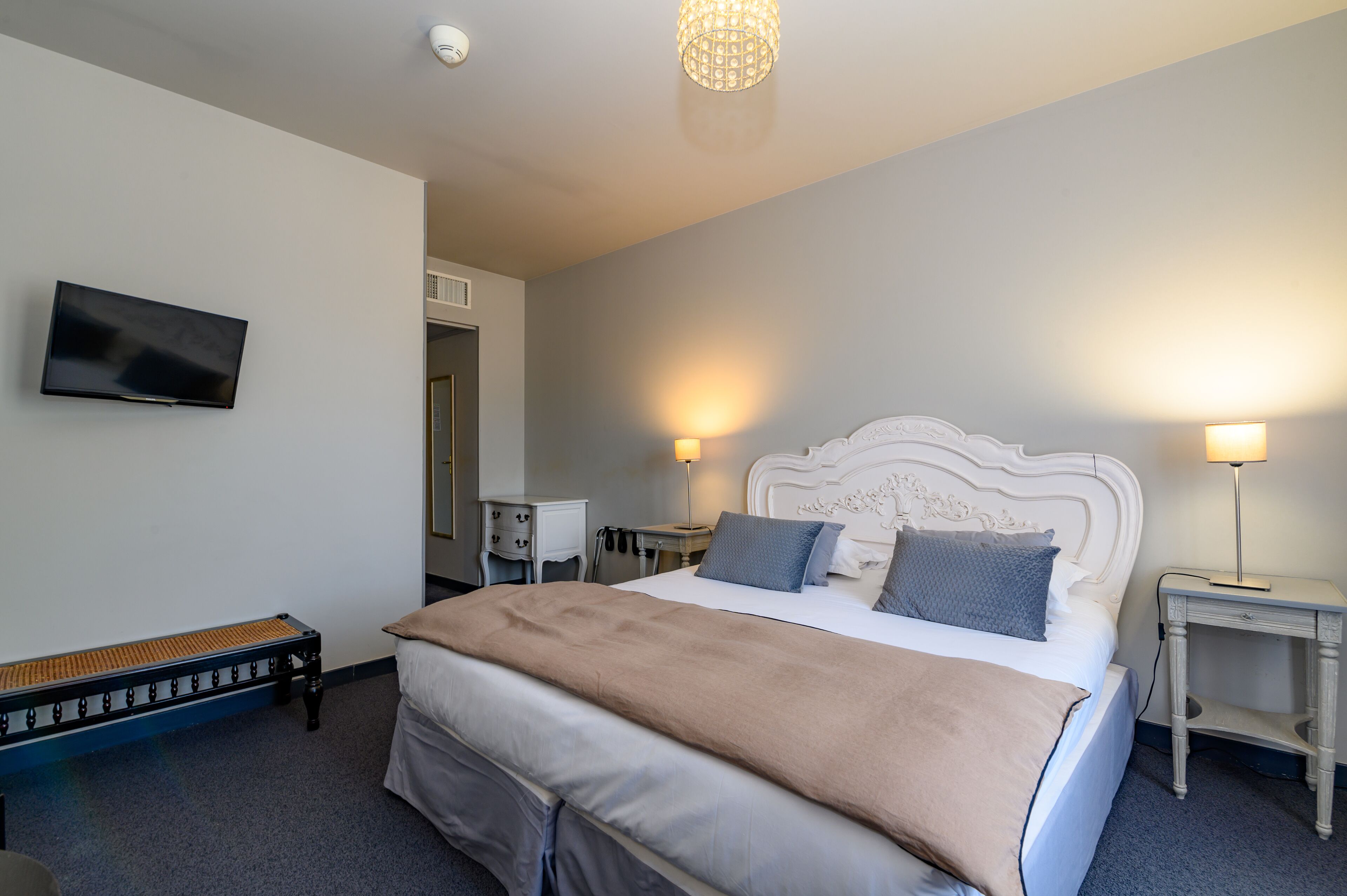 Habitación de hotel ordenada con cama, lámparas de noche, televisor y un banco.