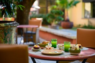 Una mesa dispuesta con alimentos para el desayuno, incluyendo tazones, platos y vasos de jugo verde, rodeada de sillas y plantas en macetas en un espacio al aire libre.
