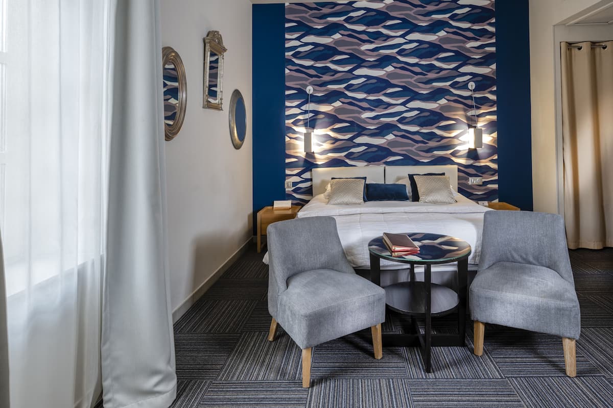 A hotel room with a bed, two armchairs, a small table, and a patterned wall.